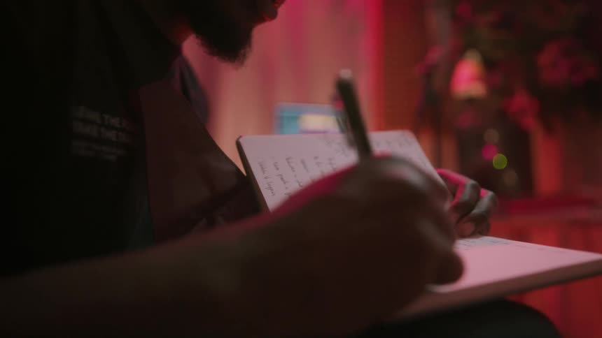 Man sitting in room lit by colored lighting writes in journal while singing