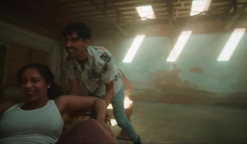 A young man pushes and twirls his friend around in a chair in an abandoned building