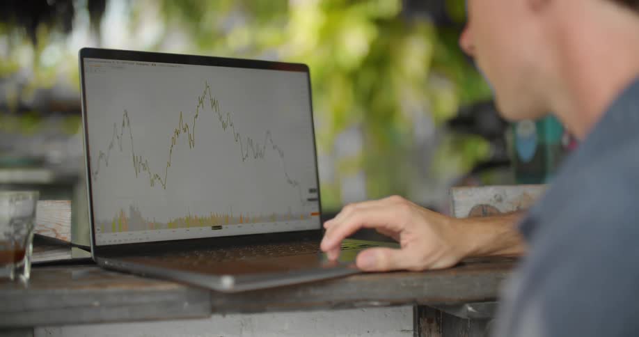 A man is sitting outside at a cafe counter looking at stock market graphs
