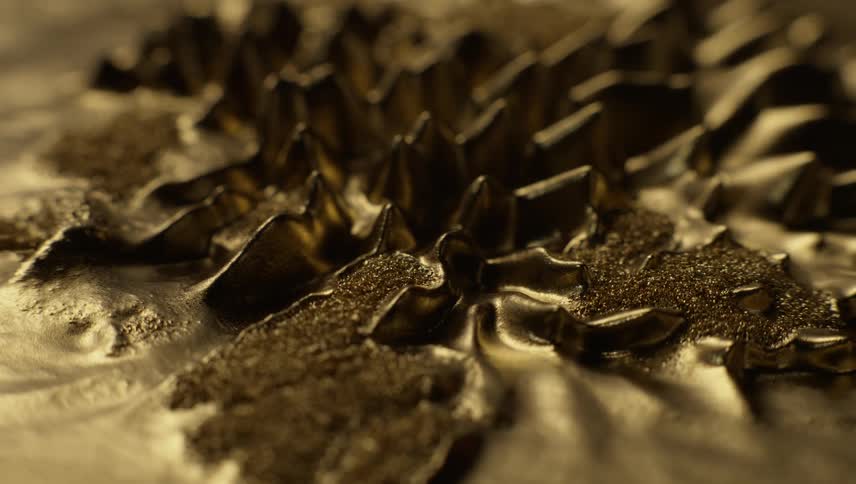 Gold liquid highly magnified moves slowly into spikes and then dissipates into a smooth liquid puddle