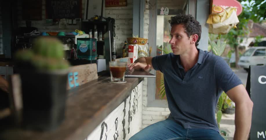 A man is sitting at the counter of an outdoor cafe typing on his laptop computer