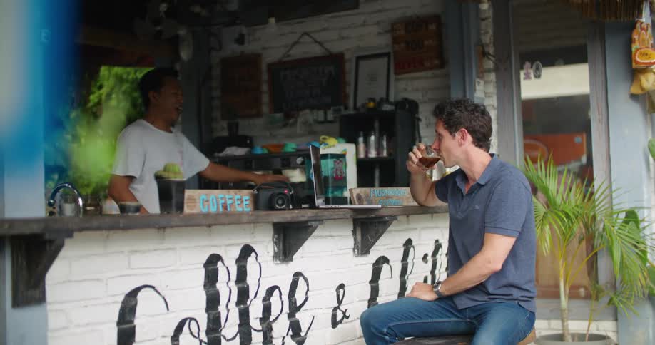 Customer sitting at open air counter accepts drink from coffee shop clerk and sips it