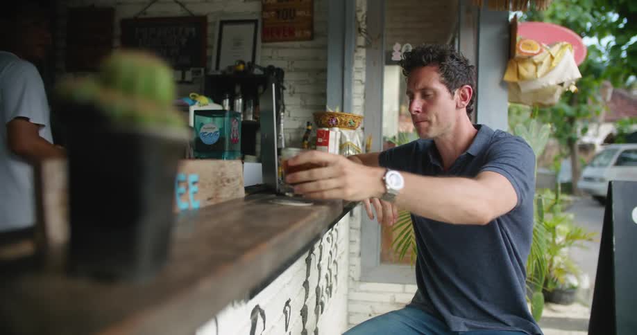 A man sits at an outdoor bar and takes a sip of his drink while someone bikes past