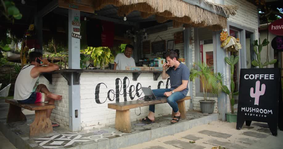A man is sitting on a bench at an outdoor cafe with his laptop computer and the man behind the counter hands him a drink