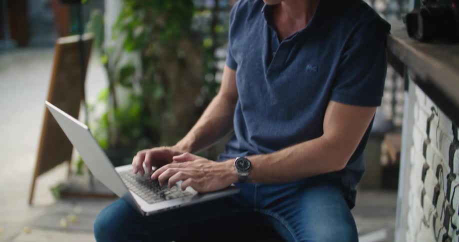 A man sitting in an open air cafe typing on a laptop with his camera on a counter