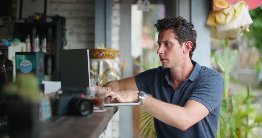 Man sitting at counter of open air caf types on laptop as others walk in the background