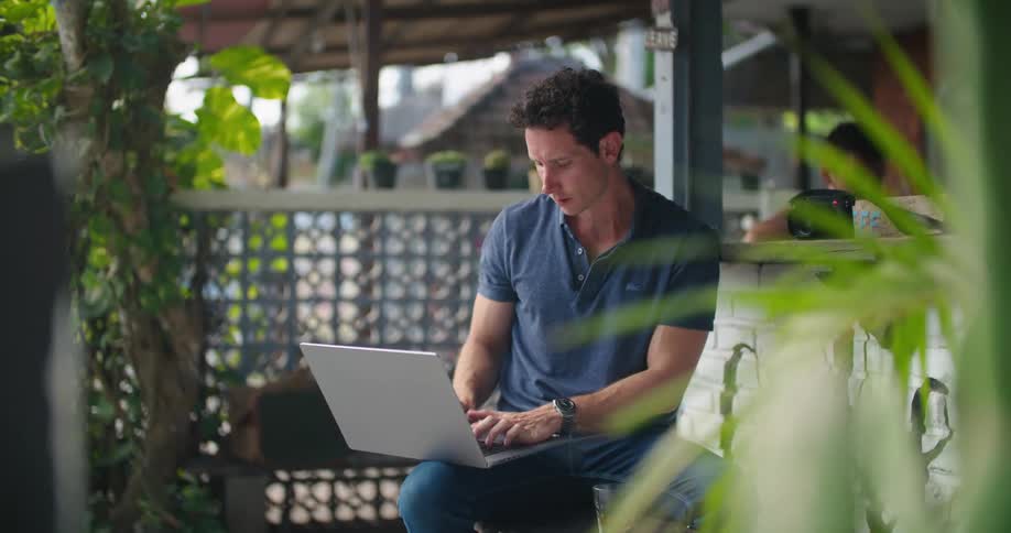A man is sitting outside on a patio typing on his laptop computer and stopping to drink from a glass sitting next to him