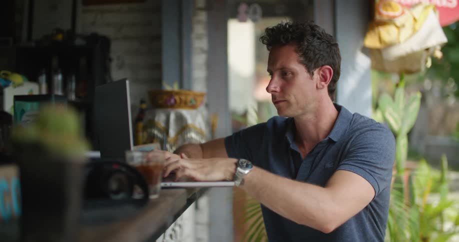 A man sips a drink while sitting at the bar typing on a laptop