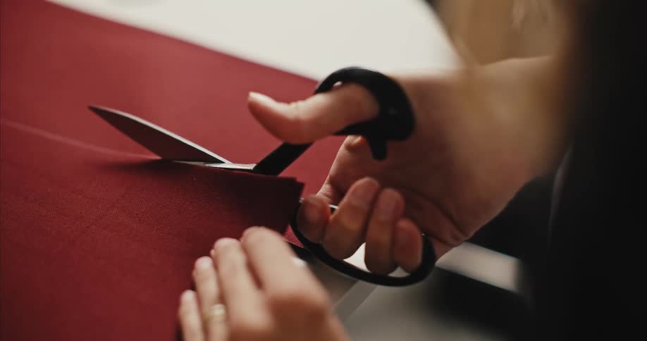 A woman is holding scissors as she cuts a maroon colored fabric