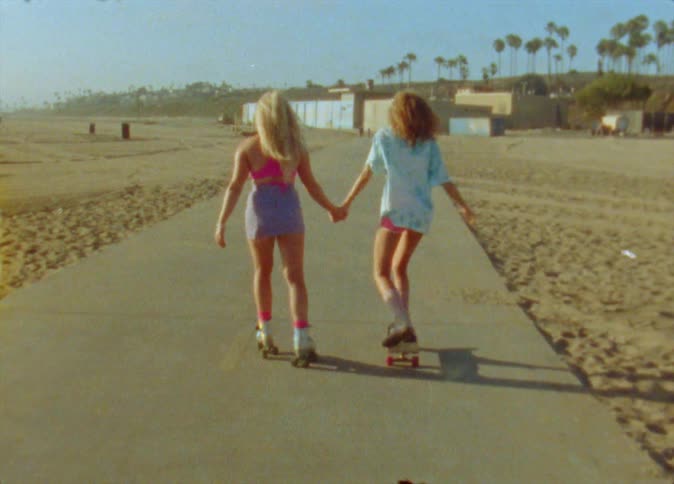 Girls with blonde straight curly hair shorts pink top and white blue shirt holding hands roller skate and skateboard on road on the beach