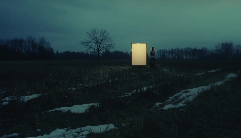 A woman stands in front of a lit up box in a snowy field at night and then takes off her cloak and steps away to turn and look at the lit up box and then walk away