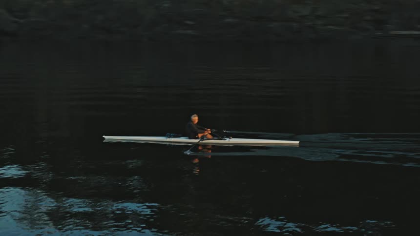 Man rows across reflective water as mist overs part of forest on shore