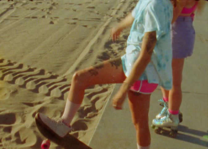 A teenage girl with roller skates stands next to another girl with a skateboard on the beach