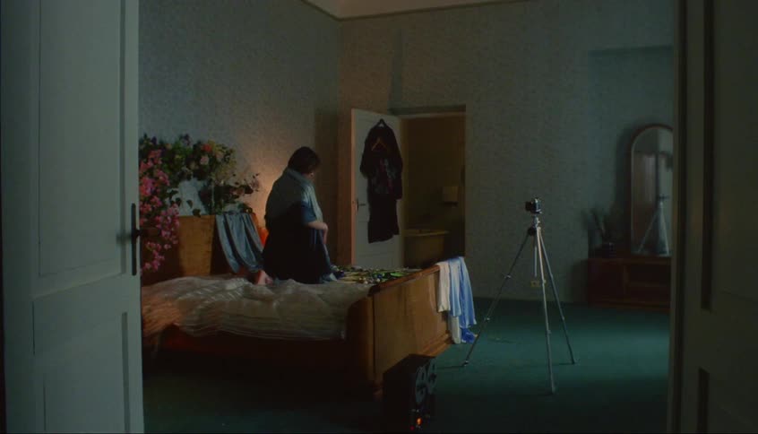 Woman with purple dress sits on bed poses for camera on tripod in bedroom
