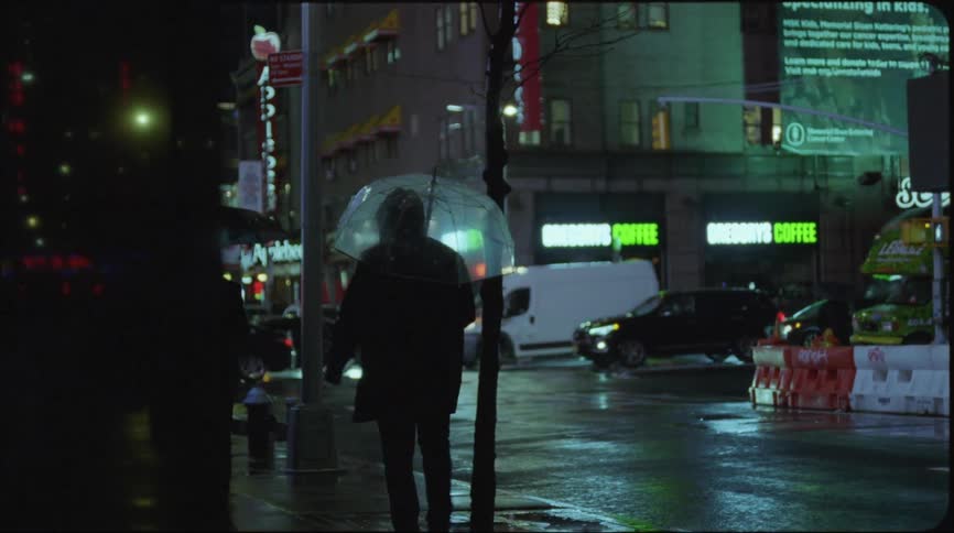 People with black coats and umbrella walk down street next to highway with cars bus and van driving while drizzling at night in the city