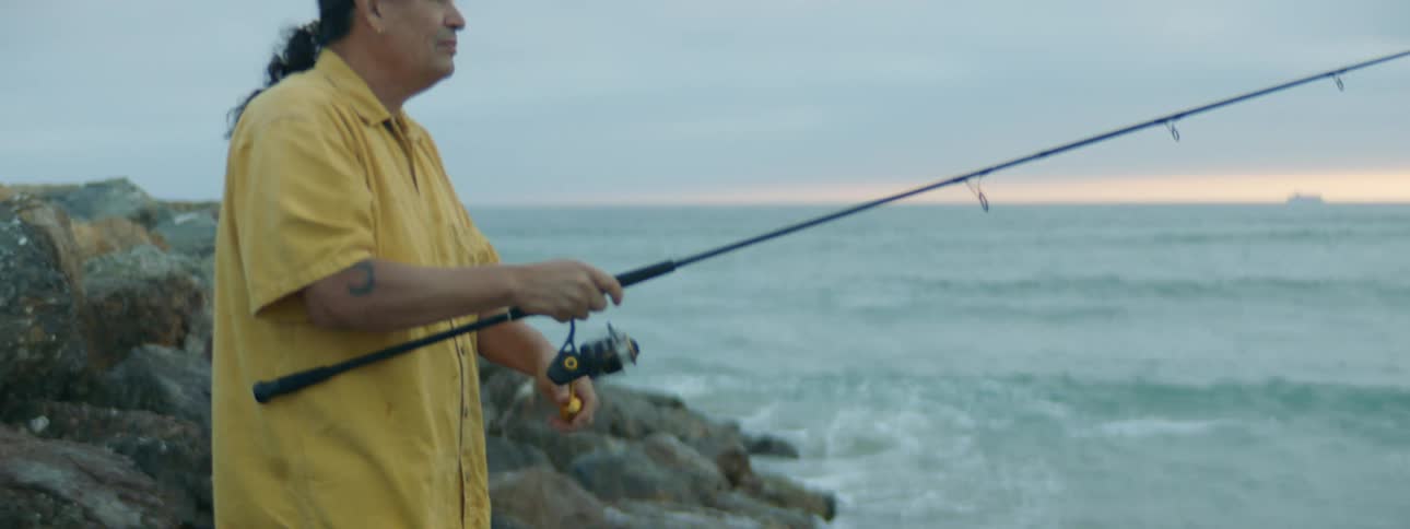 A man standing on the rocks fishing as waves break and splash up