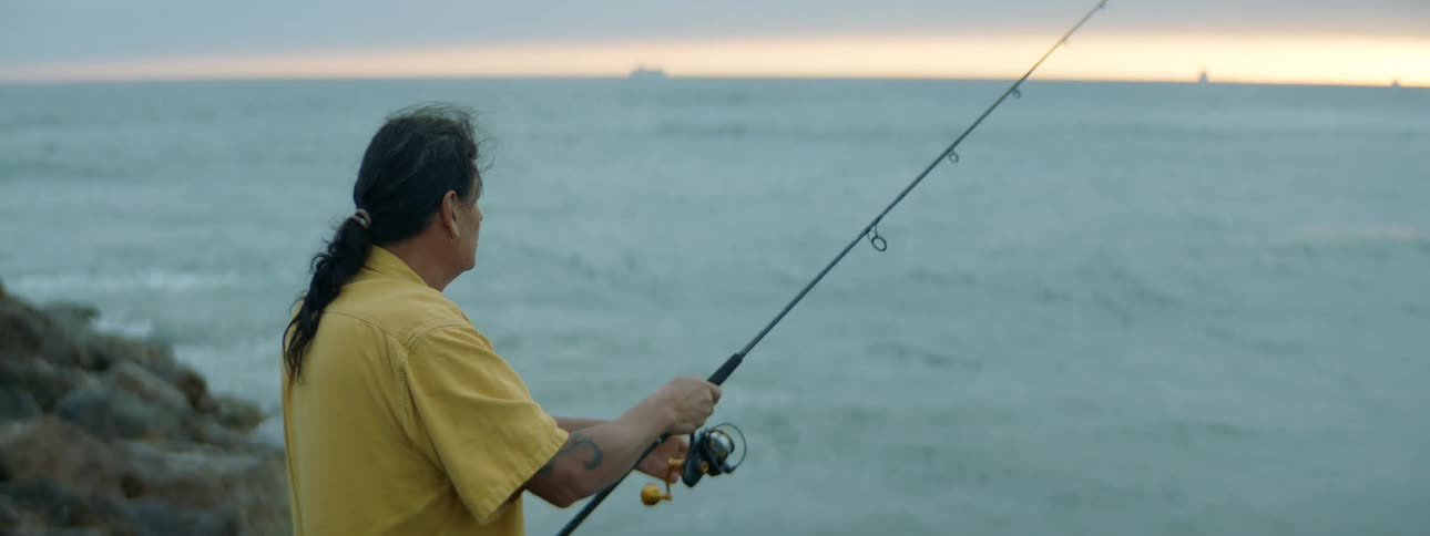 A man is standing along the coast near rocks casting the line with his fishing rod