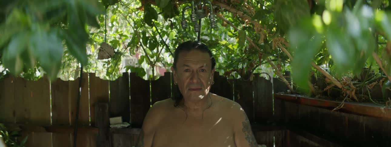 A man is sitting against a wooden fence under a tree