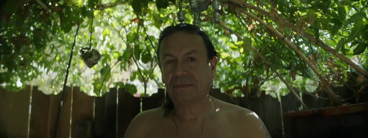 A shirtless man turns while standing in front of a fence