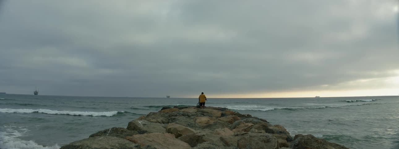 Person wearing yellow walks out onto rocky shore as boats move on horizon under an overcast sky