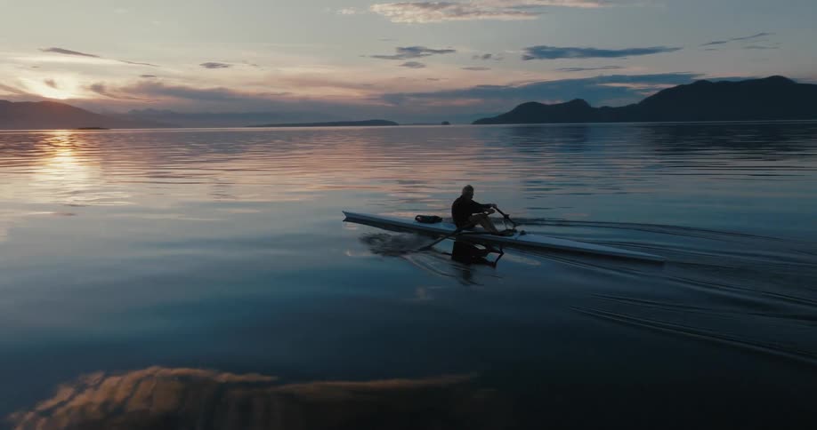 Man rows a boat using a sculls on lake next to the forest at sunset