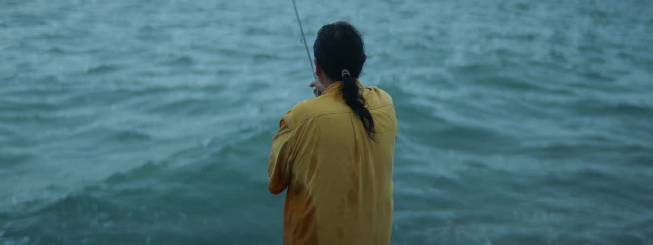 Man with ponytail and yellow shirt casts fishing pole on the coast