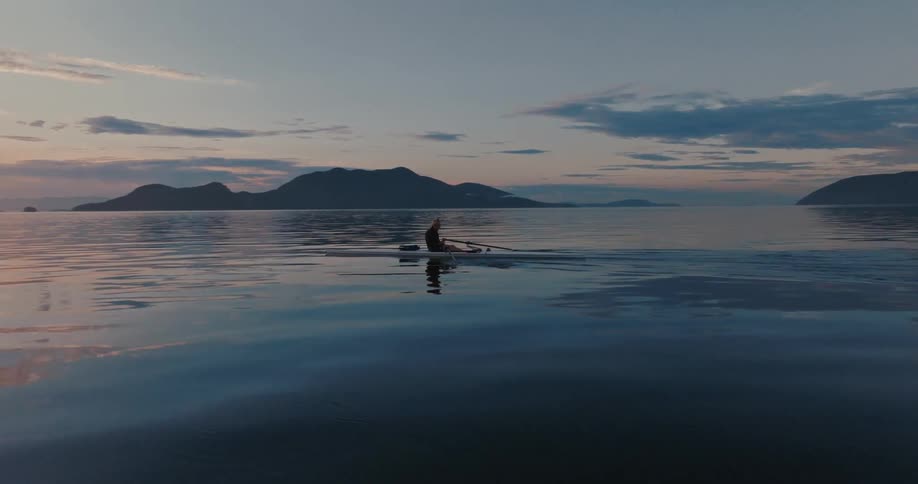 A man rows a boat in a middle of a lake with distant mountains at sunset