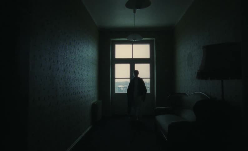 Silhouette of a woman with a blanket around her shoulders standing in front of windows and pressing her hands on the glass then walking to the couch and sitting down