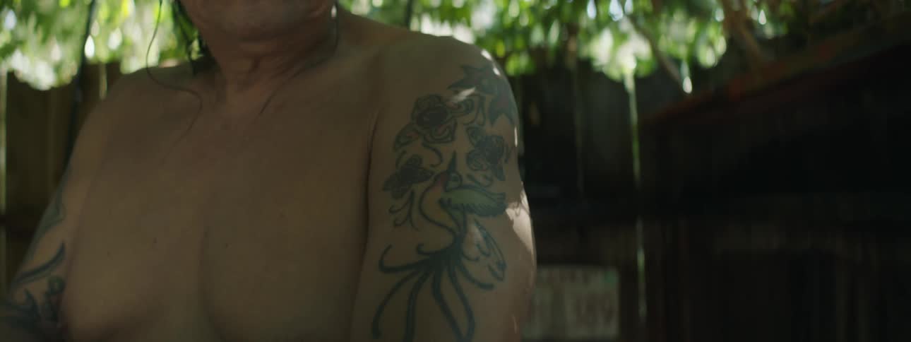 A man is standing near a wooden fence by a tree and rubbing his arm with a cloth