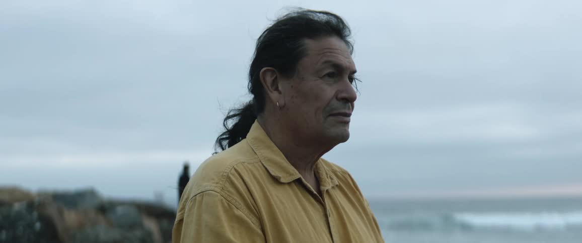 Middle aged man with ponytail and yellow shirt looks around on the coast