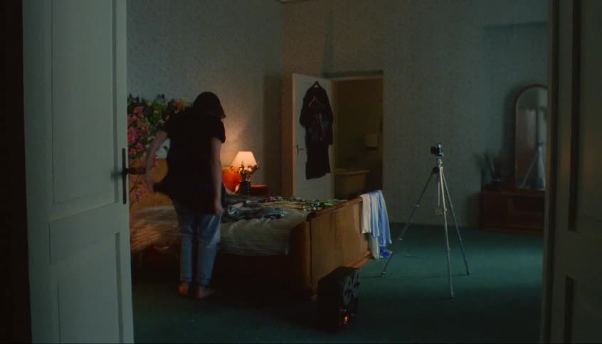 Woman putting on purple dress sits on bed poses for camera on tripod in bedroom