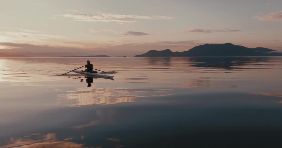Sun shines in red sky just above mountains as person rows kayak across glassy surface of water reflecting sky
