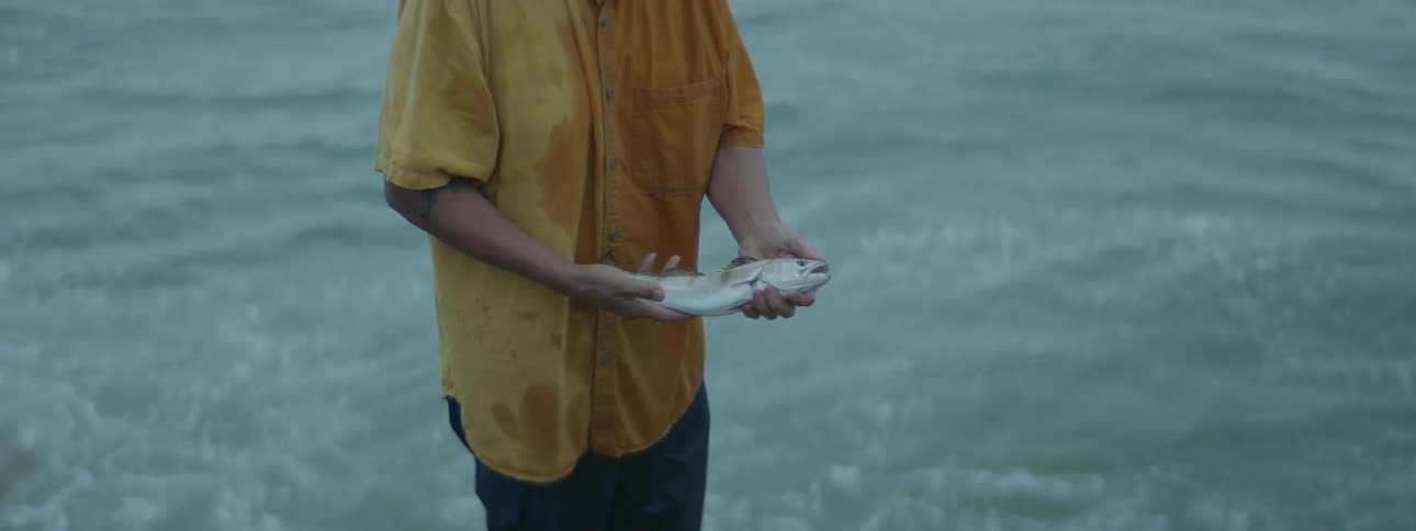 Man stands in surf in a wet shirt holding a fish