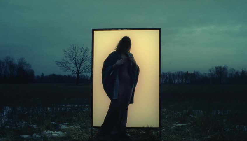 Silhouette of a woman standing in front of an illuminated framed board and she steps aways and takes off her coat