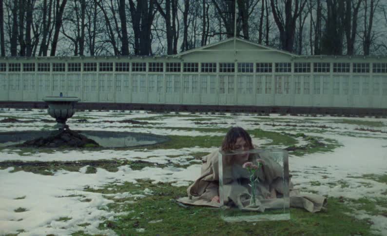 A woman is sitting outside in the grass with snow on the ground looking at a glass cube with pink flowers inside it and she picks up a crowbar and is moving it down over the cube
