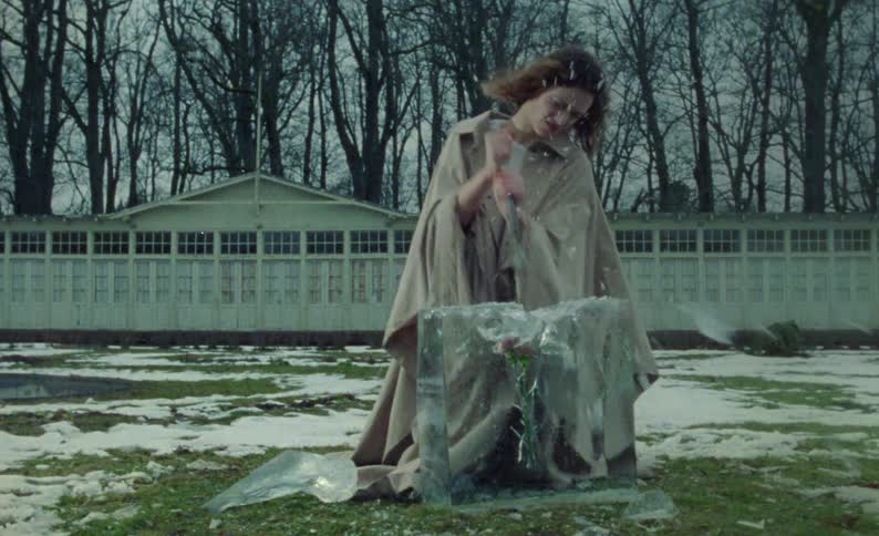A woman is outside standing over a glass cube with pink flowers inside it and breaking it with a crowbar