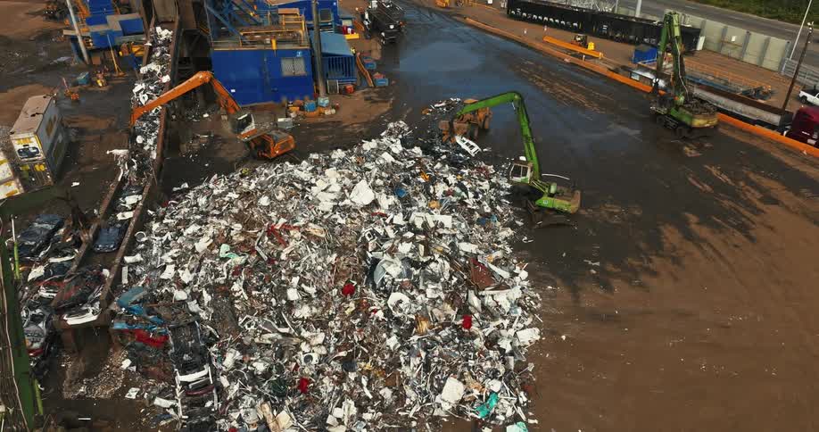 A pile of scrap is standing in a scrap yard as a junk yard crane grappler moves it and other trucks work