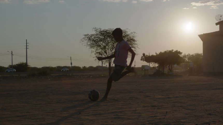 A boy is outside of his home and he kicks a ball sitting on the ground as the sun shines brightly behind him