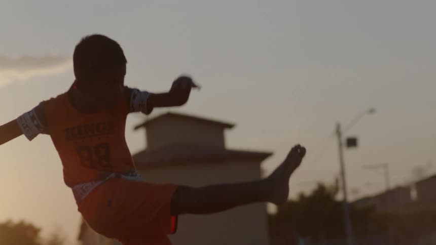 A barefoot boy kicks a soccer ball into the air while standing outdoors in the evening