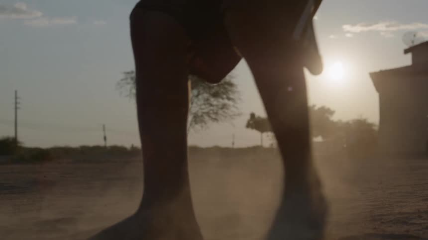The silhouette of a barefoot boy kicking a soccer ball while motorcycles travel past