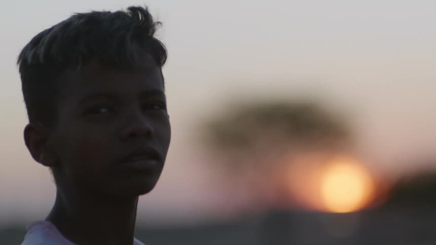 A boy standing outdoors at sunset turns his head to look into the camera