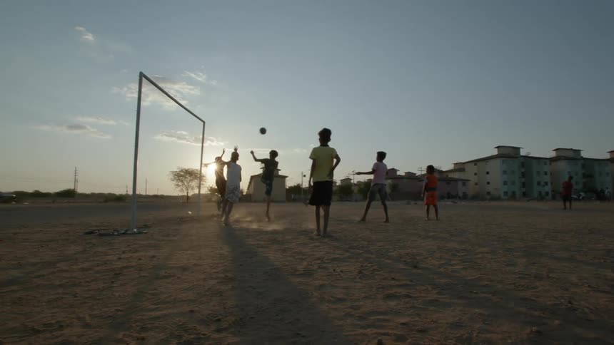 Kids play soccer in sandy field near apartment building and are silhouetted by the sun