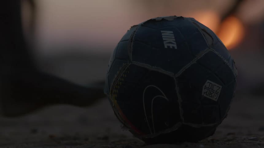 A ball sits on the ground with silhouettes of boys running in the distance and someone kicks the ball