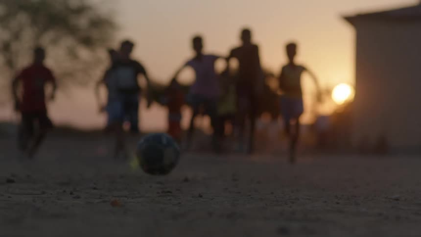 A ball sits on the ground and someone kicks it and a group of boys run forward and start playing