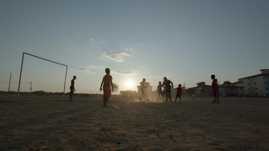 Kids partially silhouetted against the sun kick up dust as they play soccer