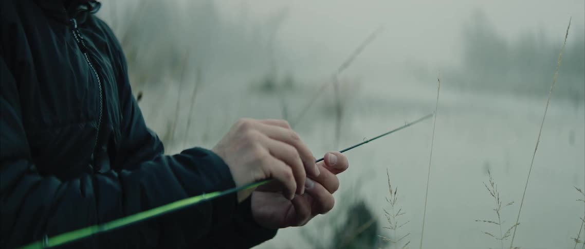 A man sits next to a river and wraps a leaf around a stick