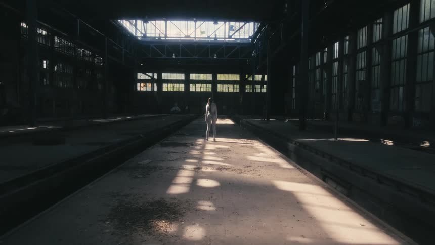 A woman is walking in an abandoned factory building and she stops to look around as sunlight shines in from the windows