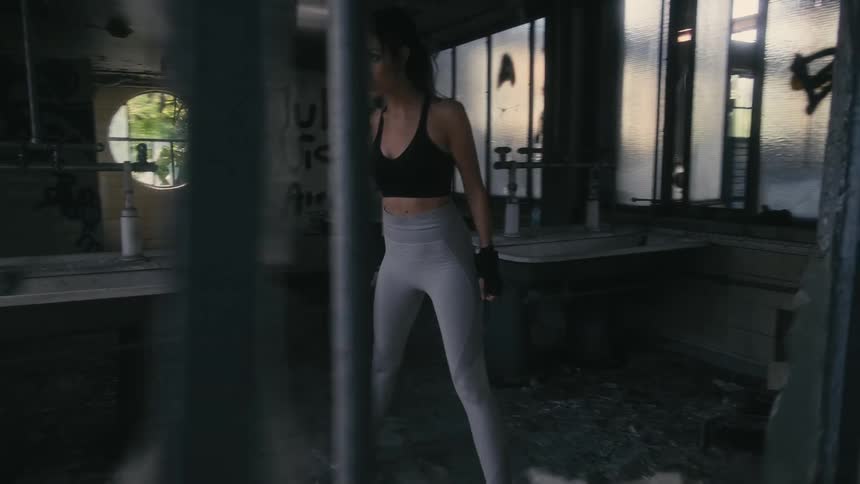 A woman is standing in an abandoned building and exercising by holding her hands together and squatting