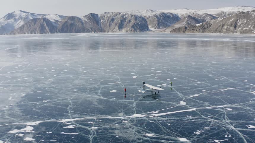 People move around airplane sitting on frozen water with snow covered mountains on the shore
