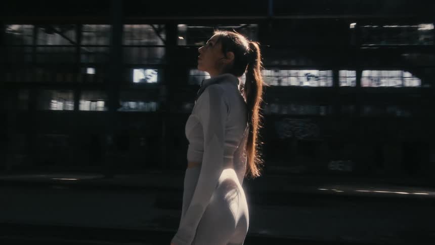 A woman is standing inside and abandoned factory building looking around as sunlight shines in from the windows